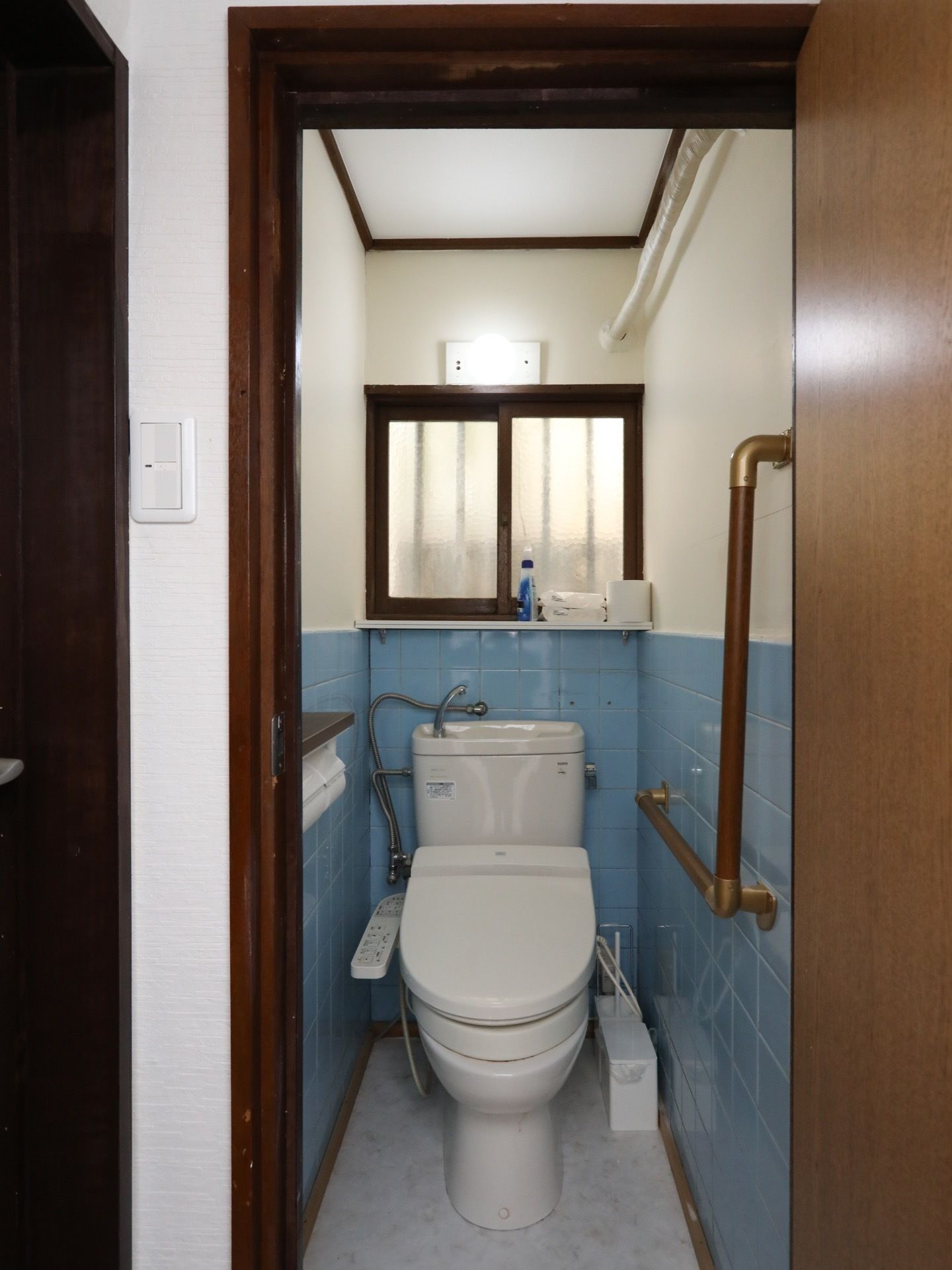 水色のタイル張りのトイレである。奥には窓があり、窓ぎわにトイレットペーパーや掃除道具が置かれている。