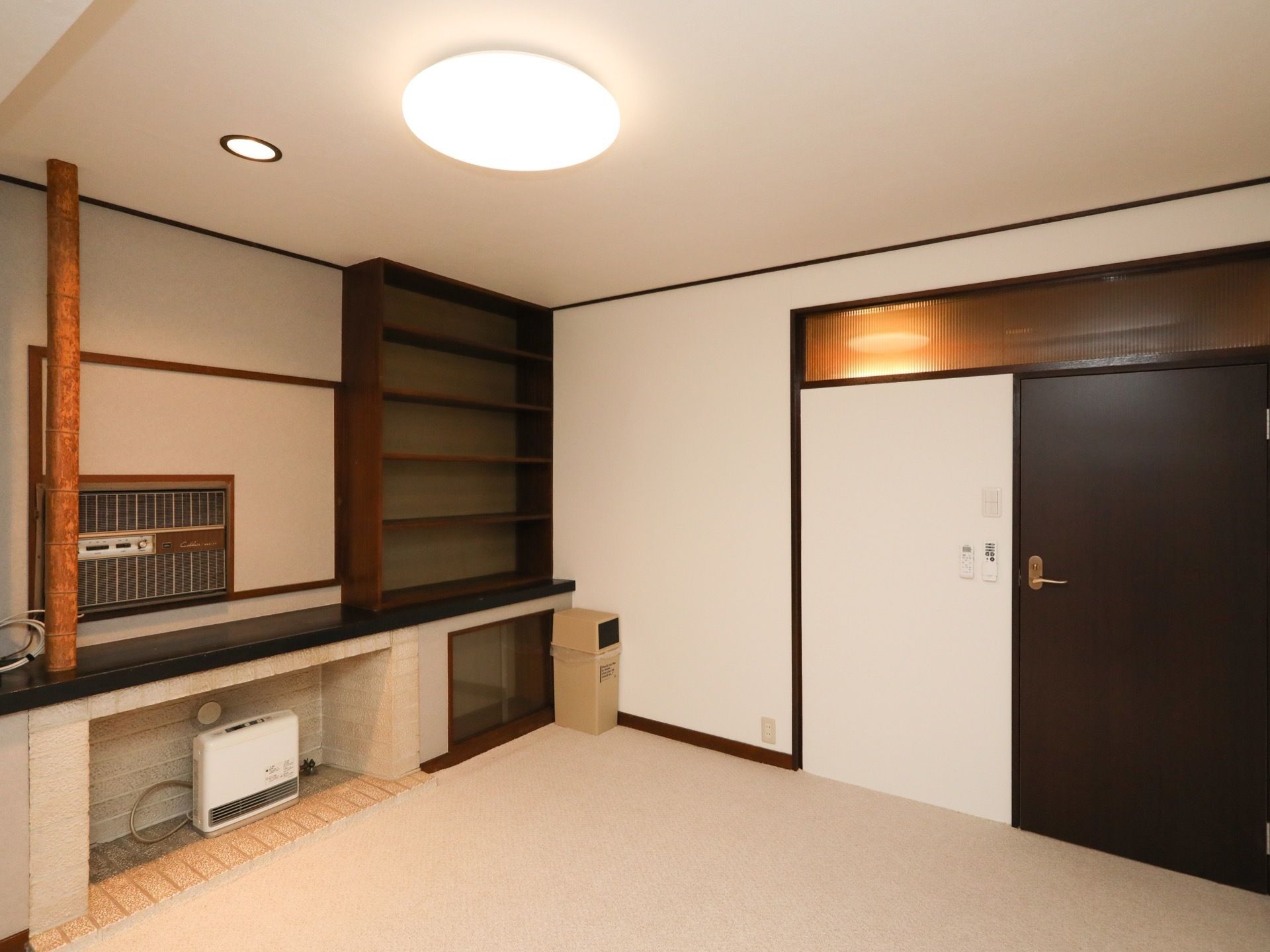 部屋の角に、壁面収納が設置されている居室である。壁側の床には暖房器具が置かれており、ゴミ箱もある。