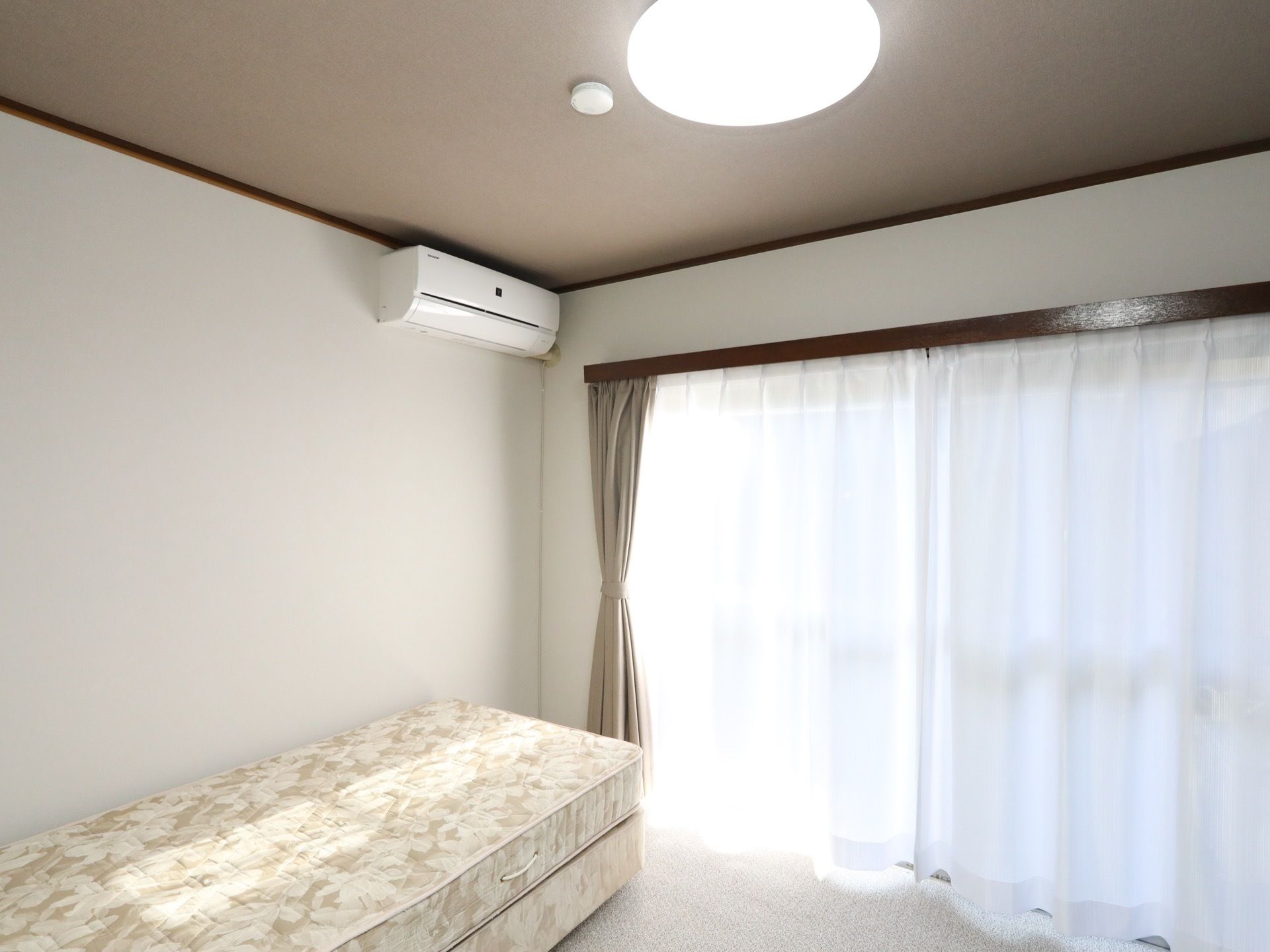 大きな窓には、白色のレースのカーテンがかけられている。また、エアコンが壁に取り付けられ、その下にベッドが置かれている。