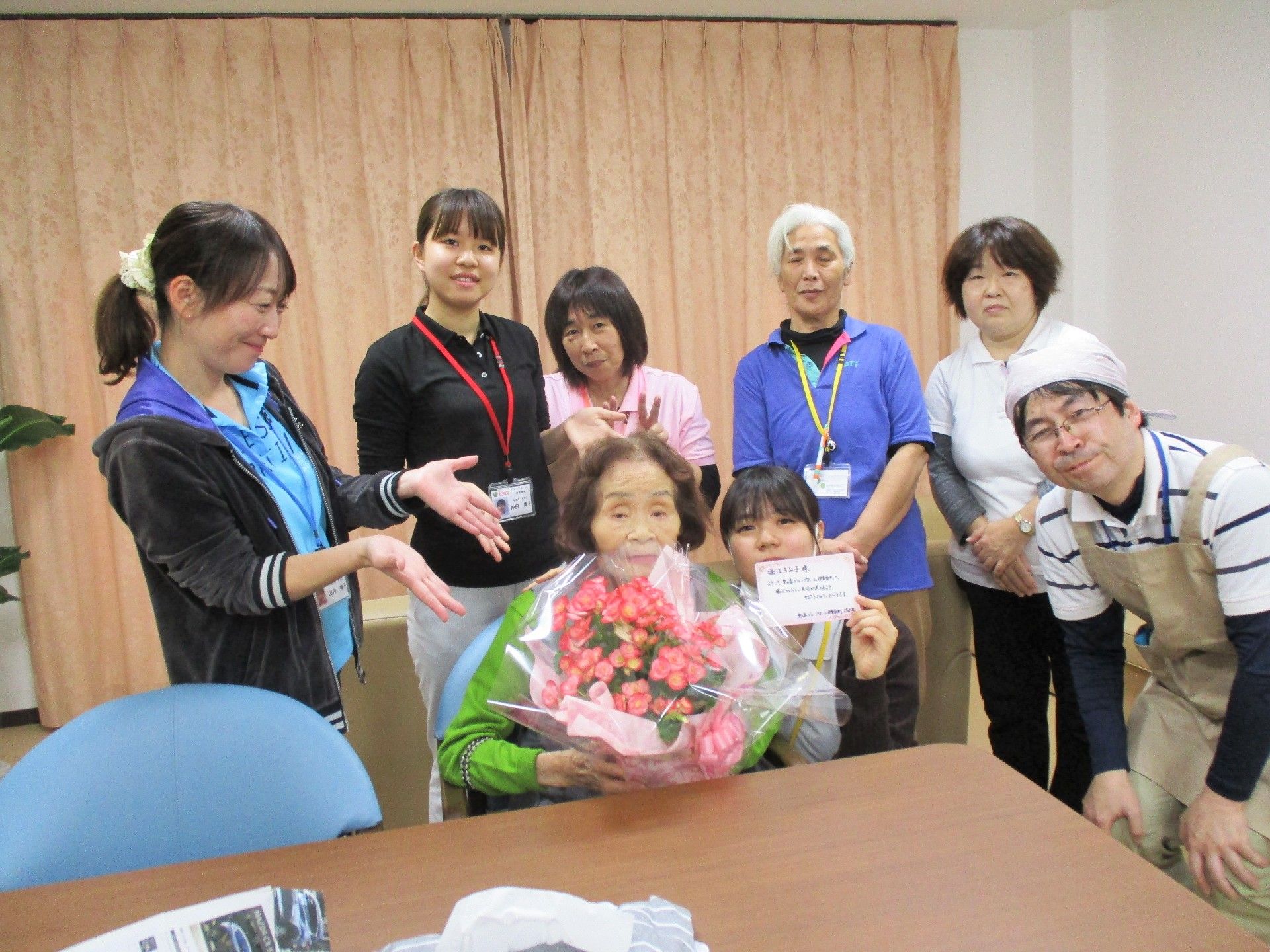 ピンクの花束を持った女性がスタッフに囲まれて記念撮影をしている様子。
