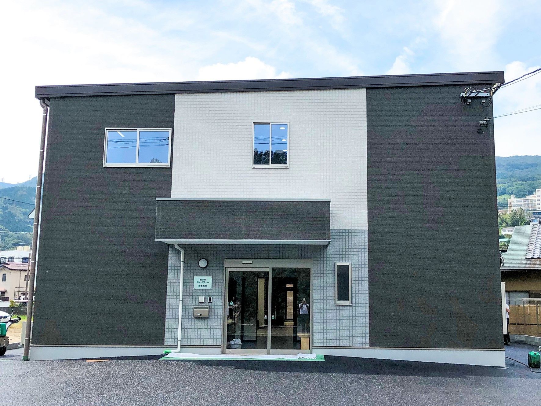 白とグレーの2階建ての建物。玄関横には駐車スペースがあり、周辺には住宅地がある。