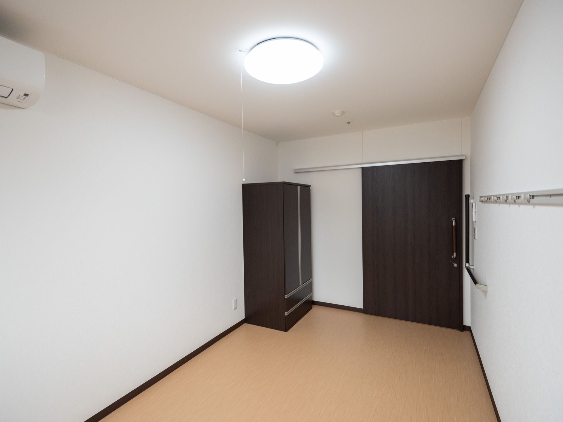 清潔感のある白が基調の居室である。照明はLEDライトで、洋服ダンス、エアコン、Ｌ字型の手すりが完備されている。