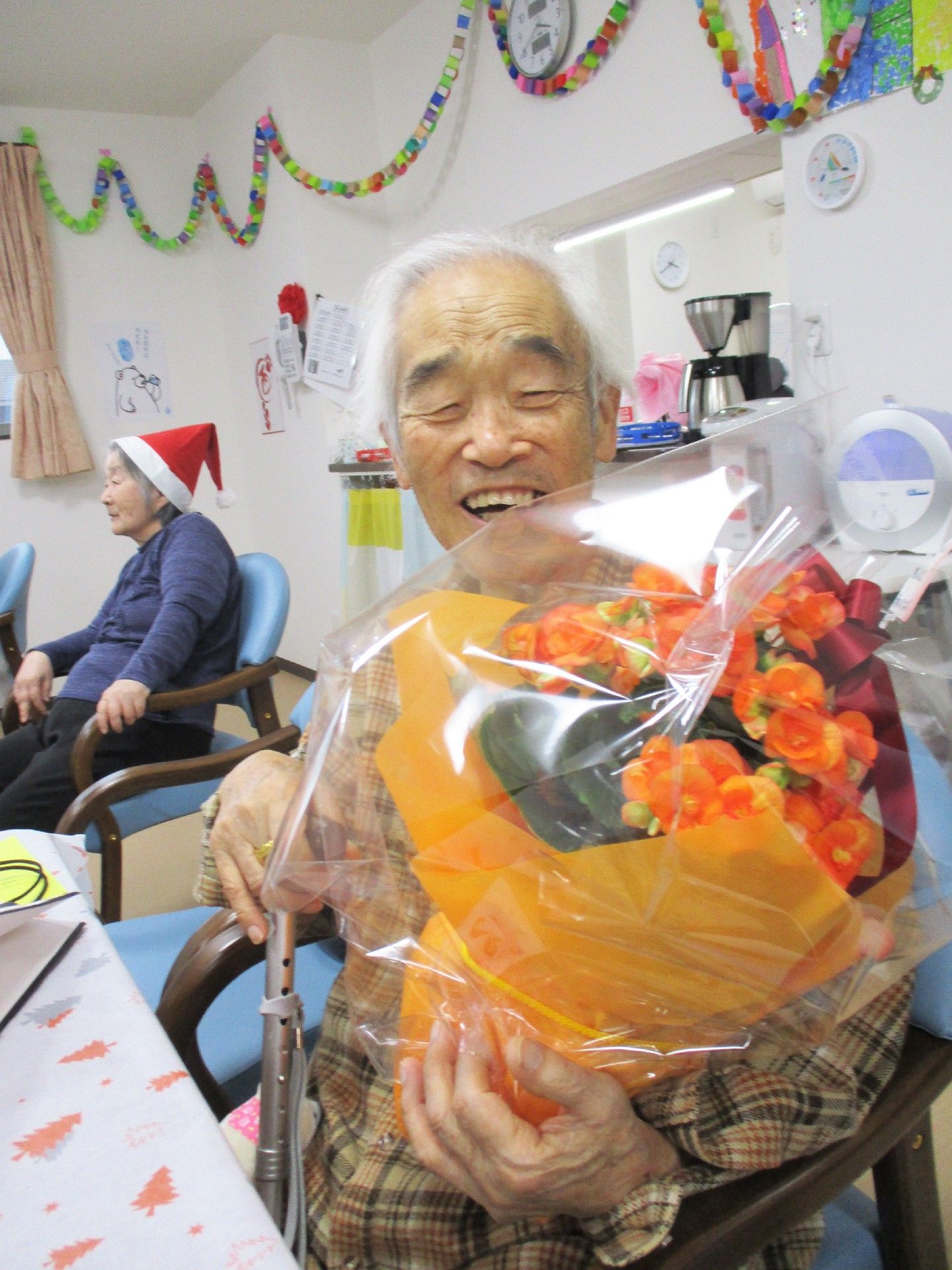オレンジの花束もらって満面の笑顔を浮かべる入居者。右手には杖を持っている。