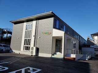 グレー系の外壁にはホーム名が書かれている。2階建ての建物の玄関には手すり付きのスロープもあり、駐車場もある。