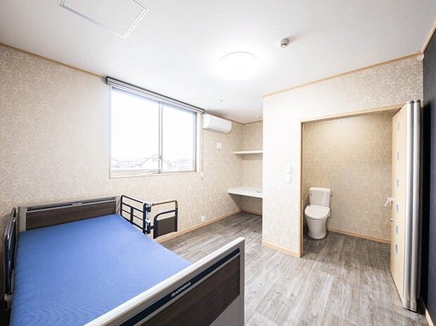 広いトイレが付いたフローリングの部屋。手前にはブルーの介護ベッドがあり、奥にはエアコンや棚が付いている。