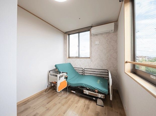 グリーンの介護ベッドが窓際に設置されたフローリングの部屋にはエアコンが設置されている。