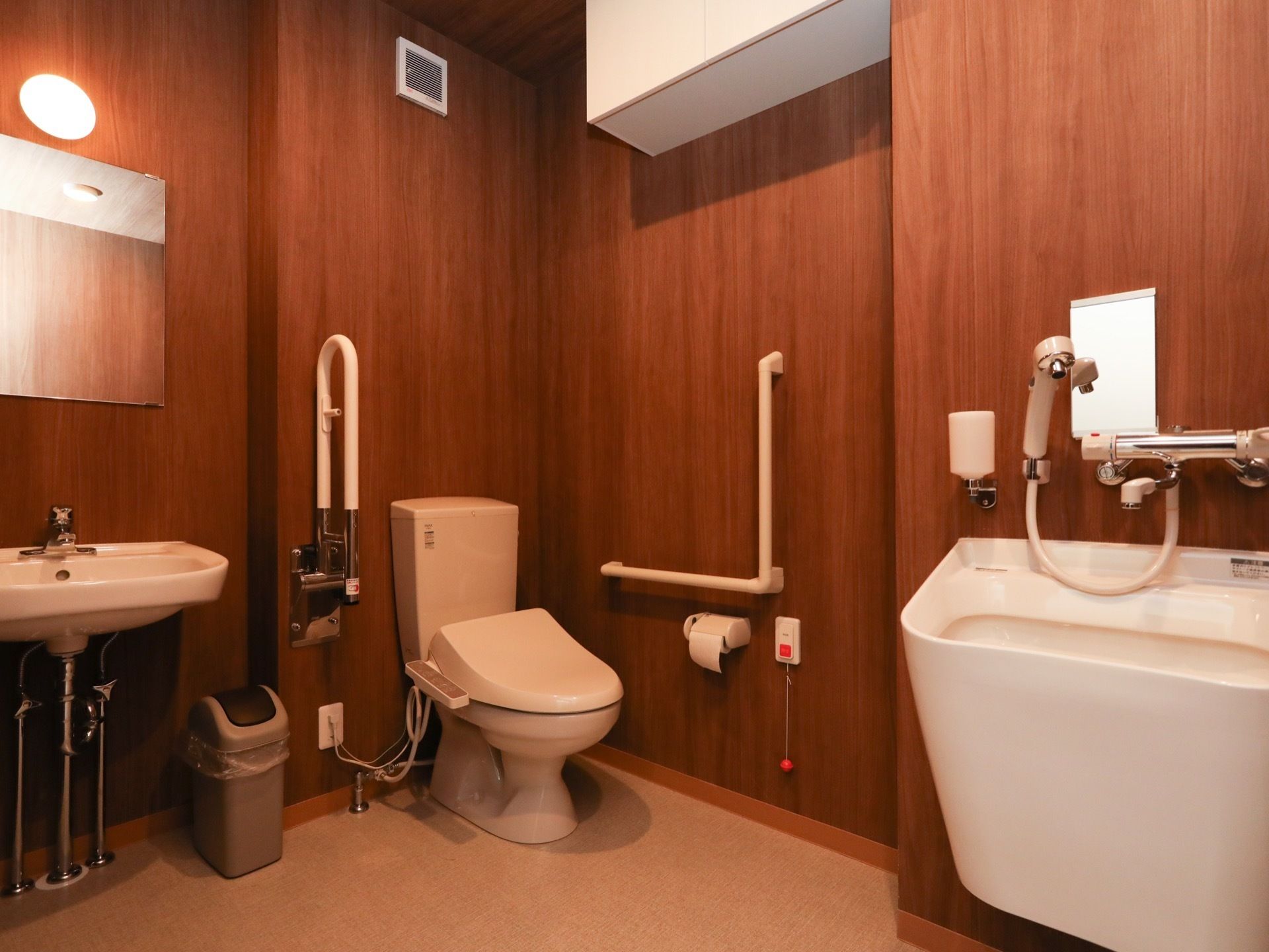 茶色い木目調の壁紙に囲まれた空間に、洋式便器とオストメイト対応のトイレが設置されている。左側には手洗器と鏡がある。