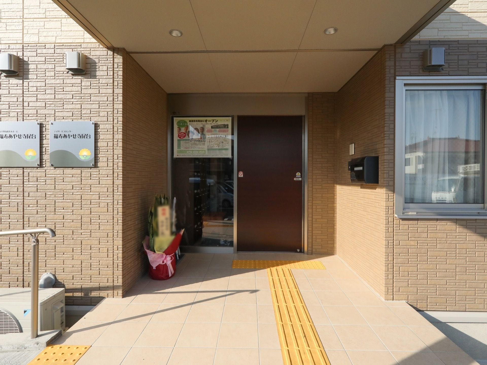 扉の右側にある壁に郵便受けが設置されている。左側の外壁に、施設名称のはいった金属製の表示板が取り付けてあり、手前に手すりが見える。