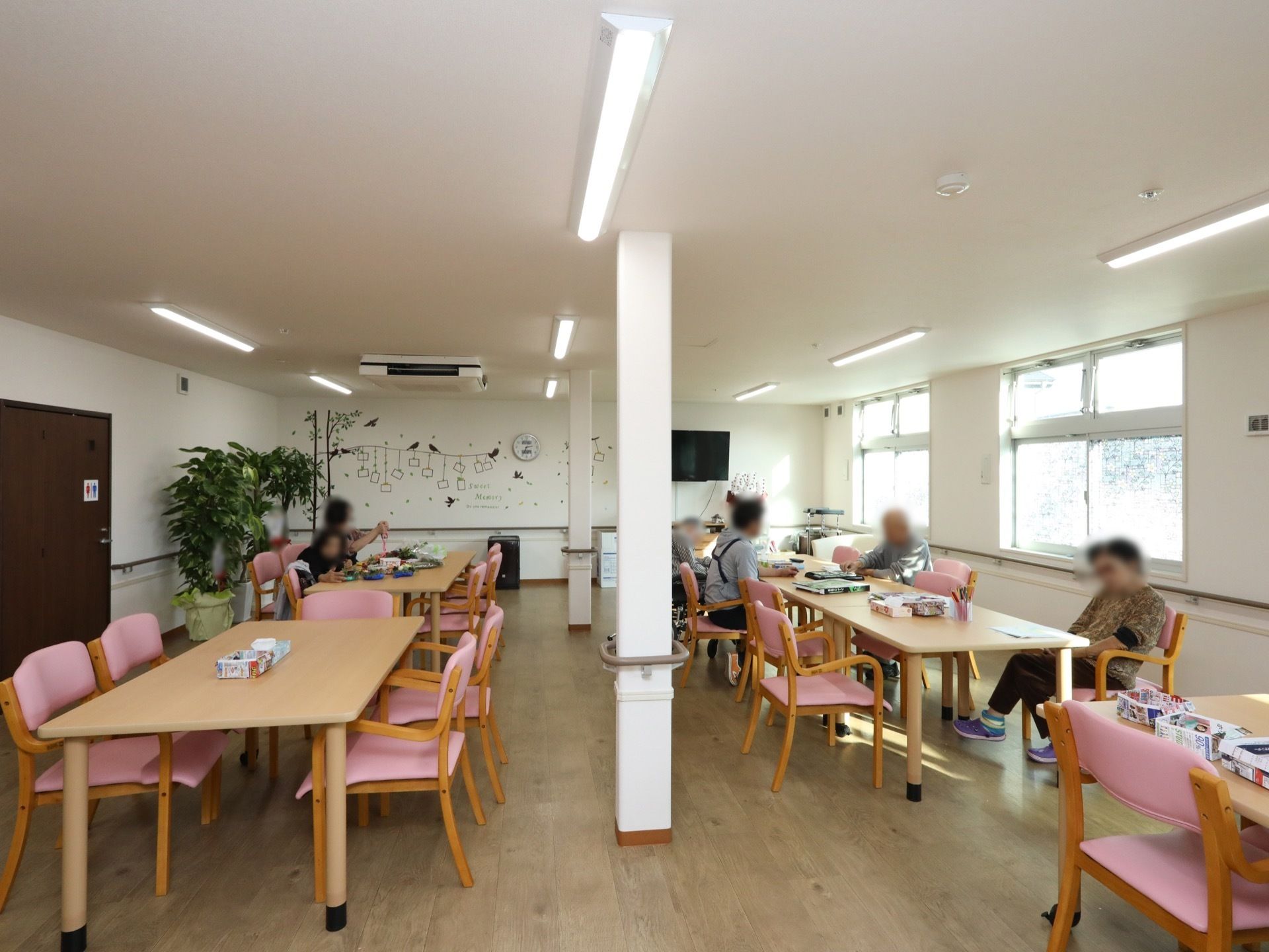 複数のテーブルや椅子が並べられている広間で、数人の入居者様が過ごしている様子。左側に茶色いトイレのドアがある。