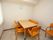 窓がある部屋に壁掛け時計が取り付けてある。テーブルを囲むように、背もたれと座面がオレンジ色の介護用椅子が配置されている。