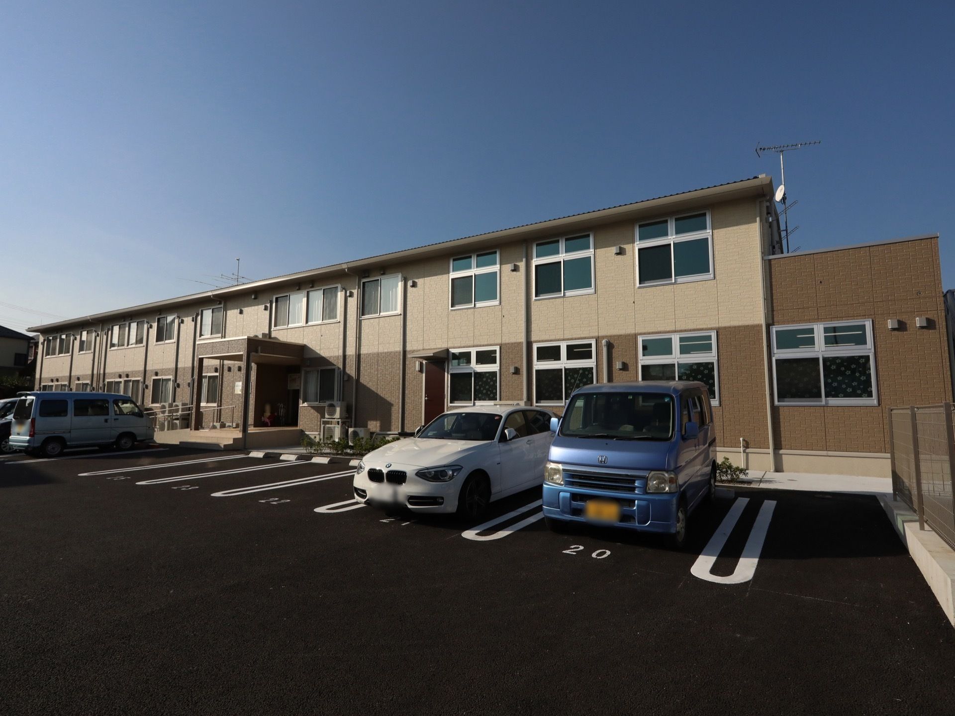 茶色い外壁が施された２階建ての施設が建っている。建物の前面はアスファルトで舗装されており、白線で１台分ずつ駐車スペースが明確に示してある。