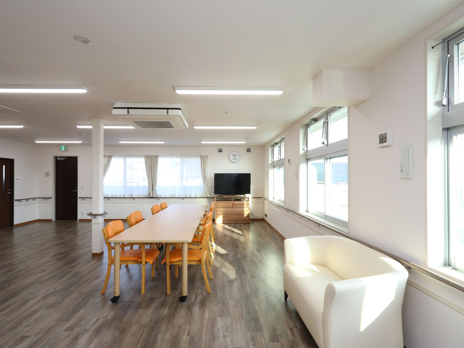 部屋の角にテレビがあり、椅子が８脚並んだテーブルと白いソファが置いてある共有スペース