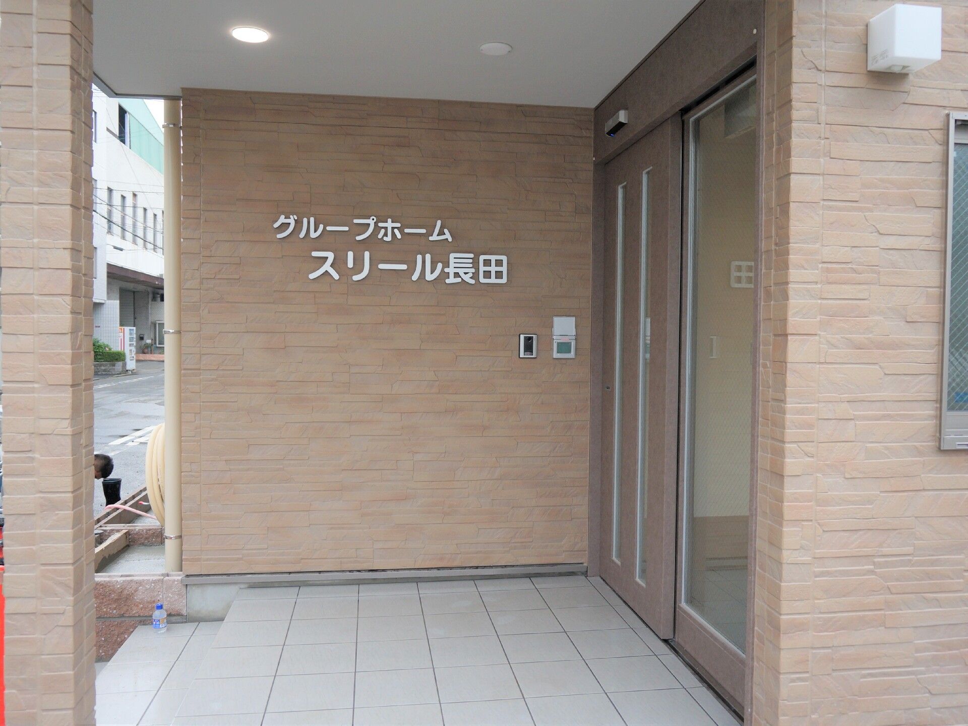 黄色いカーディガンを着た高齢女性が車いすに座り、そばにはピンクのエプロンを付けた若い女性スタッフが笑顔でいる。