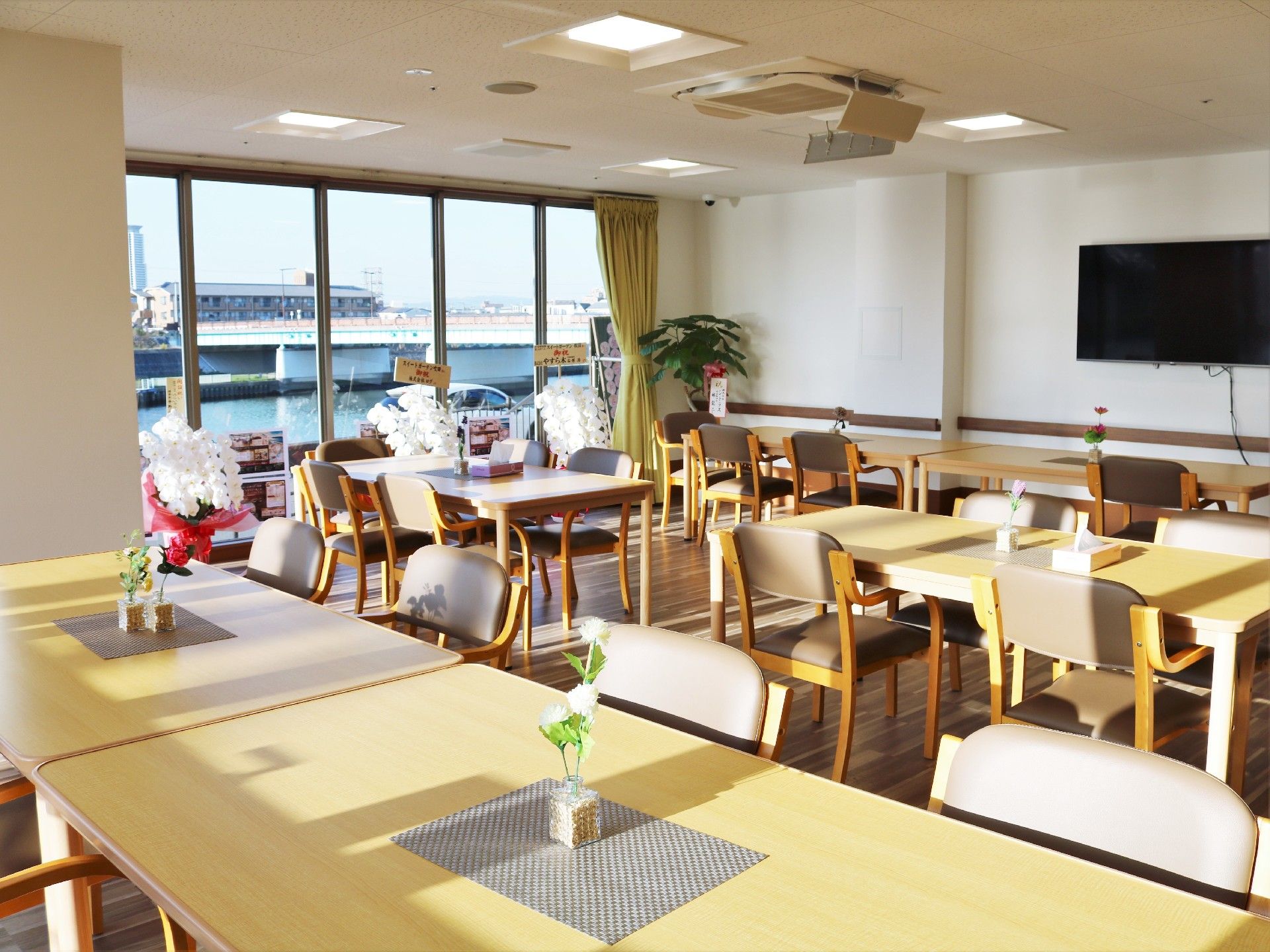 花が飾られたテーブルセットが複数置かれた食堂には大型の壁掛けテレビや観葉植物もある。