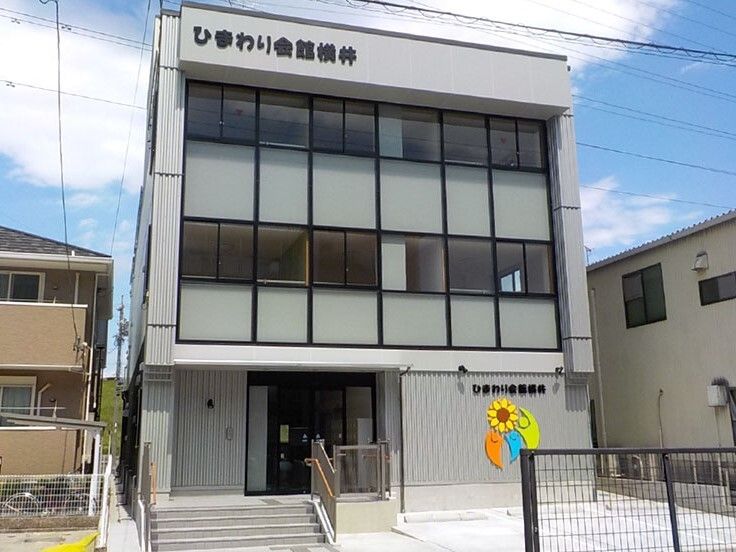 3階建てのグレー系の建物の前には駐車場があり、外壁にはホーム名が書かれ、シンボルマークが描かれている。