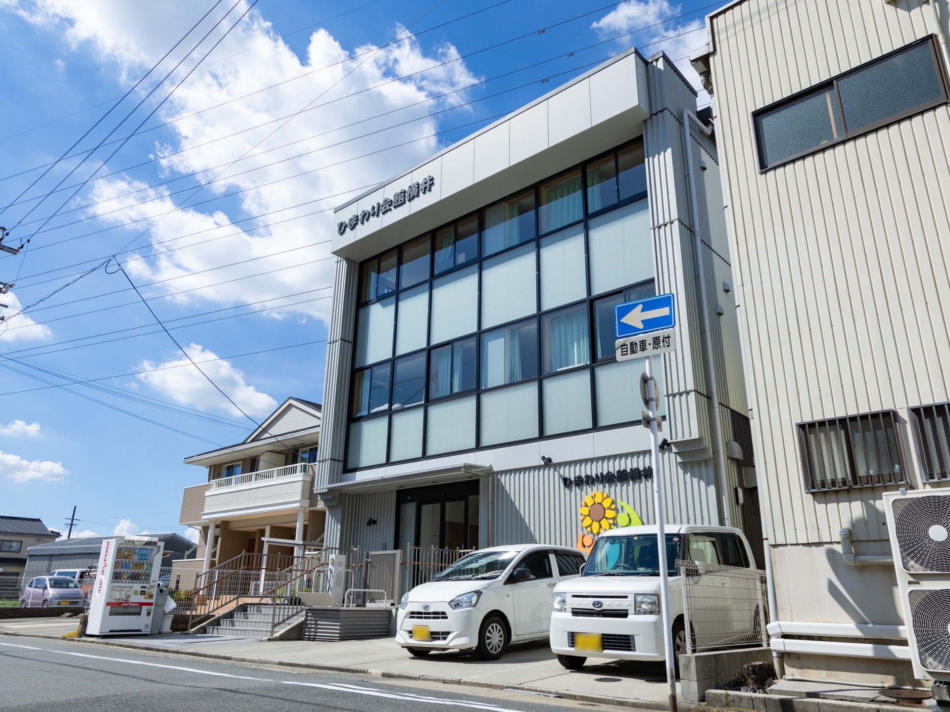 手すり付きの階段が設置された玄関の横には駐車場があり、3階建ての建物にはたくさんの窓が付いている。