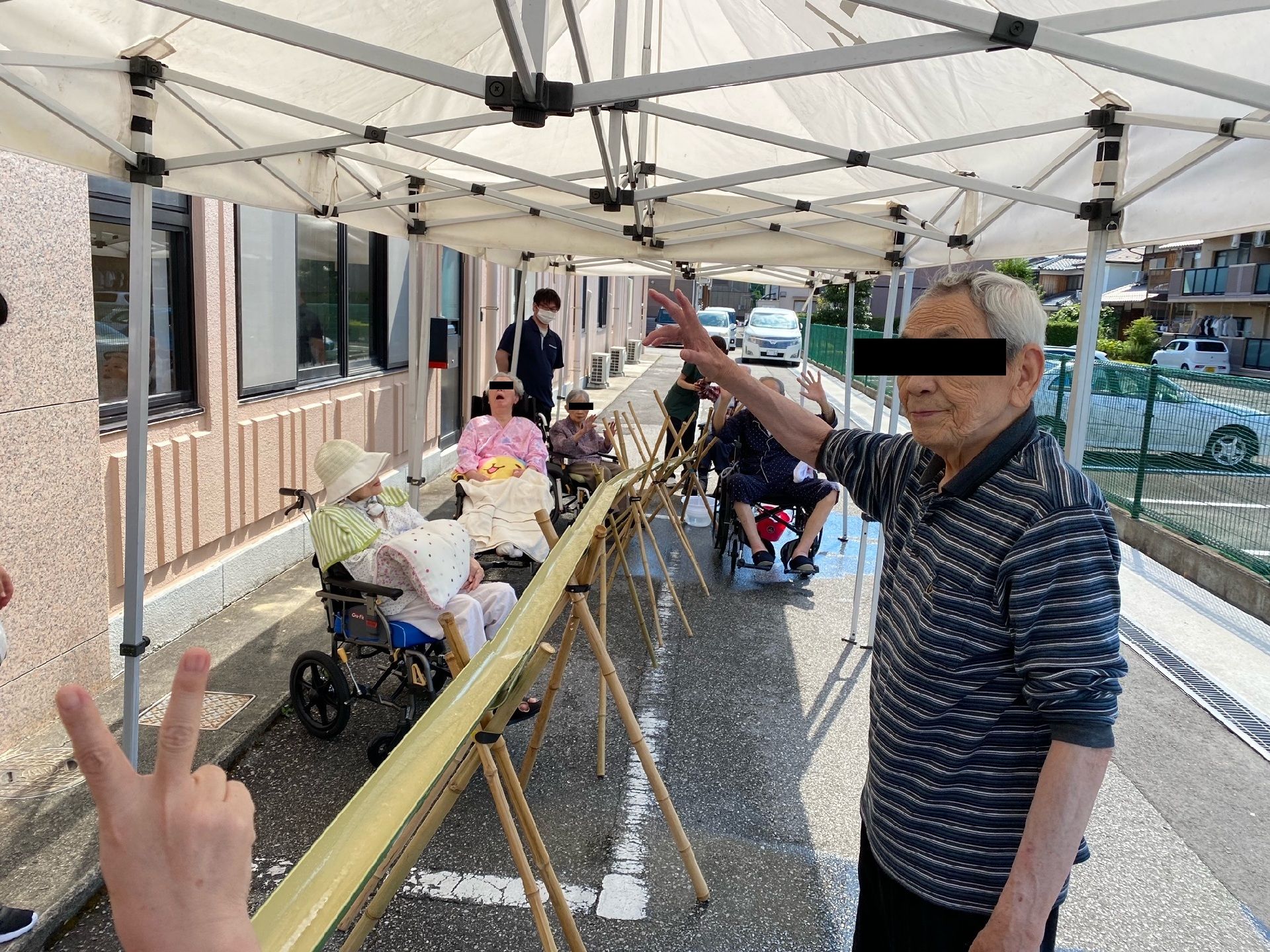 テントの下、建物の敷地内で、竹からそうめんを流し、食べている。
