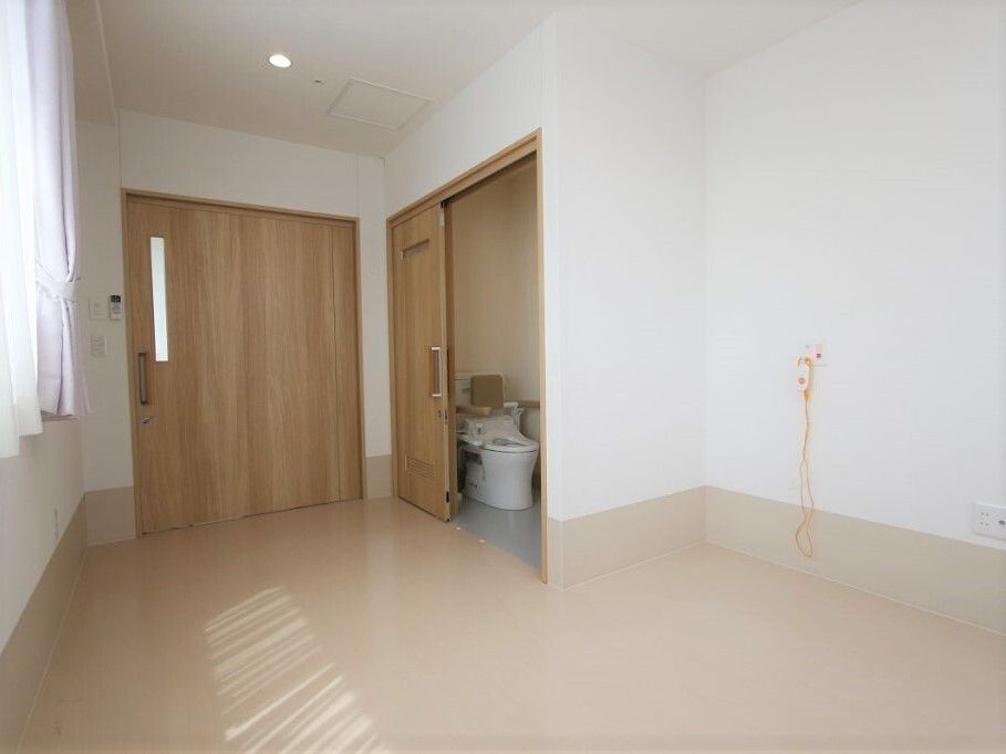 引き戸が付いたトイレが設置された個室の部屋。出入り口には大きな引き戸が付いていて、窓にはカーテンとレースカーテンが付いているが家具はない。