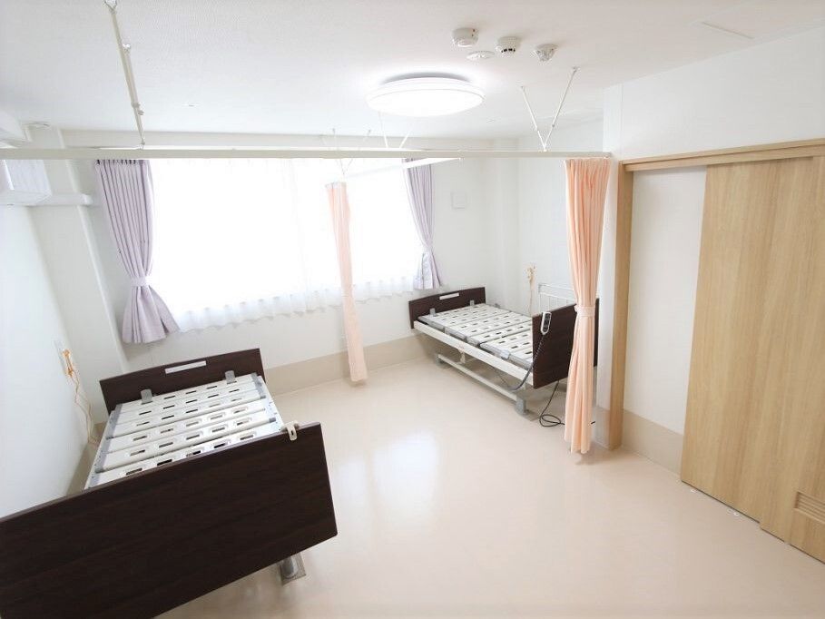 二つの介護ベッドが置かれ、日当たりのいい広い部屋。ベッドとベッドの間には仕切り用のピンクのカーテンが付いている。居室のドアはスライド式。