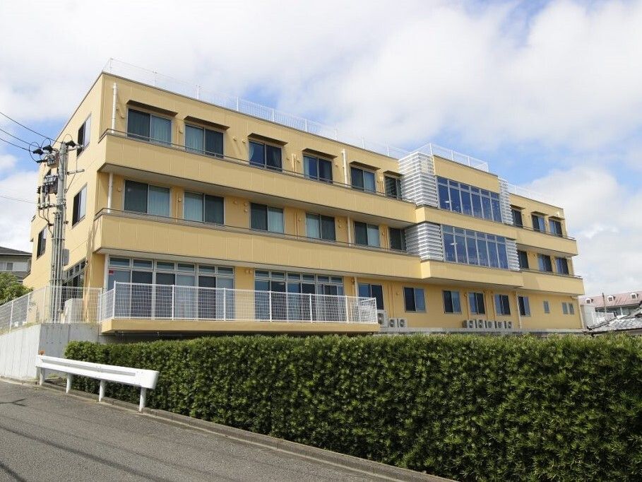 黄色がかった３階建ての建物は重厚感があり、ベランダも各フロアに設置され、緑が周辺に植えられている。周りには住宅もある。