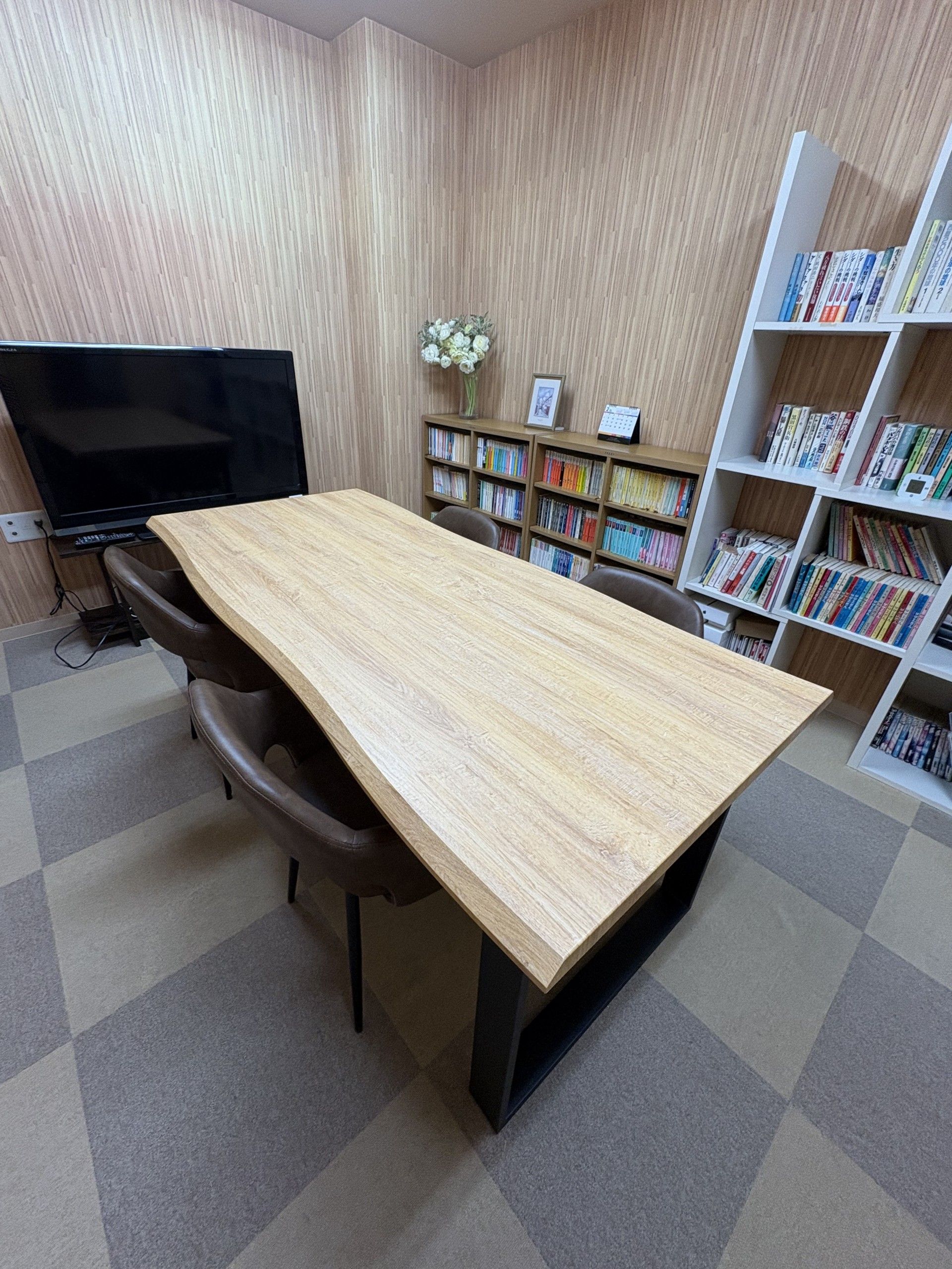 木製の長テーブルの上に、本が2冊重ねて置かれている。壁際には本棚があり、単行本や雑誌が並べられている。