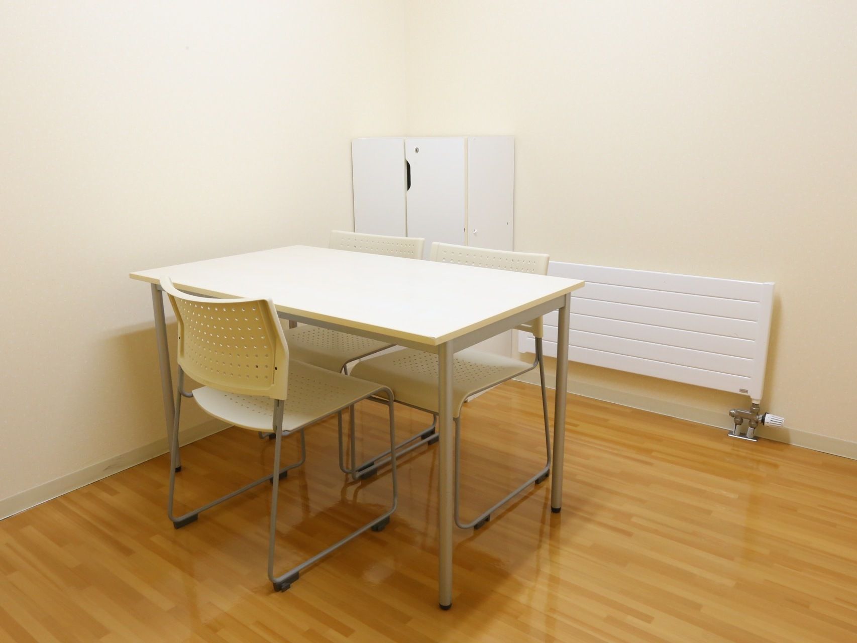 フローリングの部屋に、白い机と椅子が3脚置かれている。壁際には白い木製の収納ケースが設置されている。