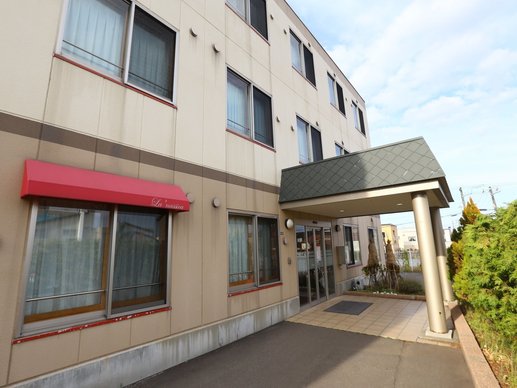 コンクリートで舗装されたアプローチが玄関に続いている。玄関部分には屋根がついている。周りには木々が植えられている。