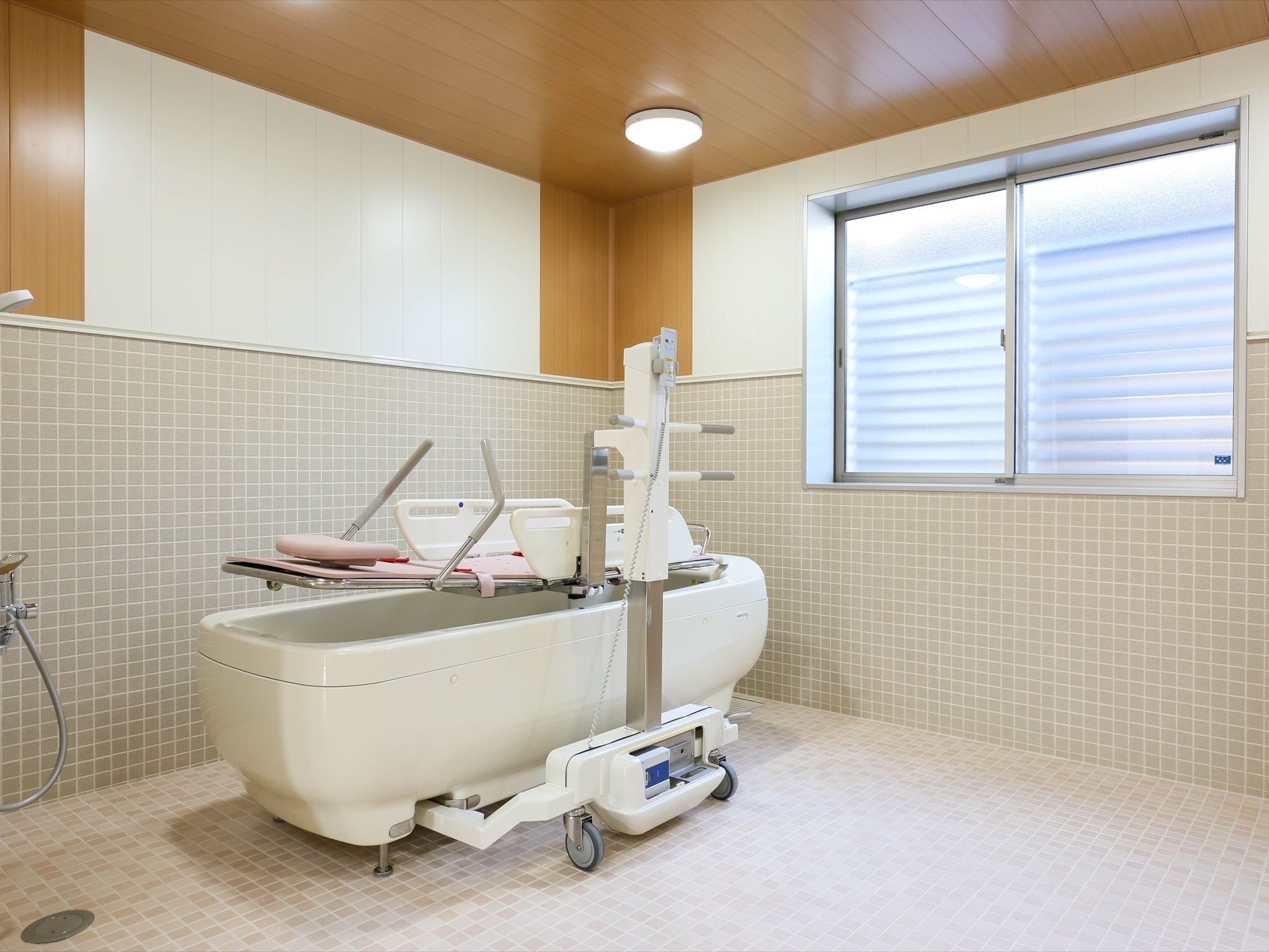 広い浴室にストレッチャー付きの介護浴槽が設置されている。床や壁は小さな灰色のタイル張りになっている。