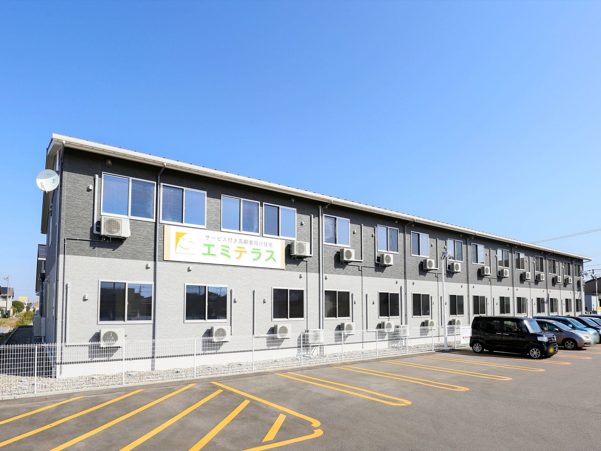 施設は２階建てで、１階部分は明るいグレー、２階部分は暗いグレーの外壁になっている。施設の横には駐車場がある。