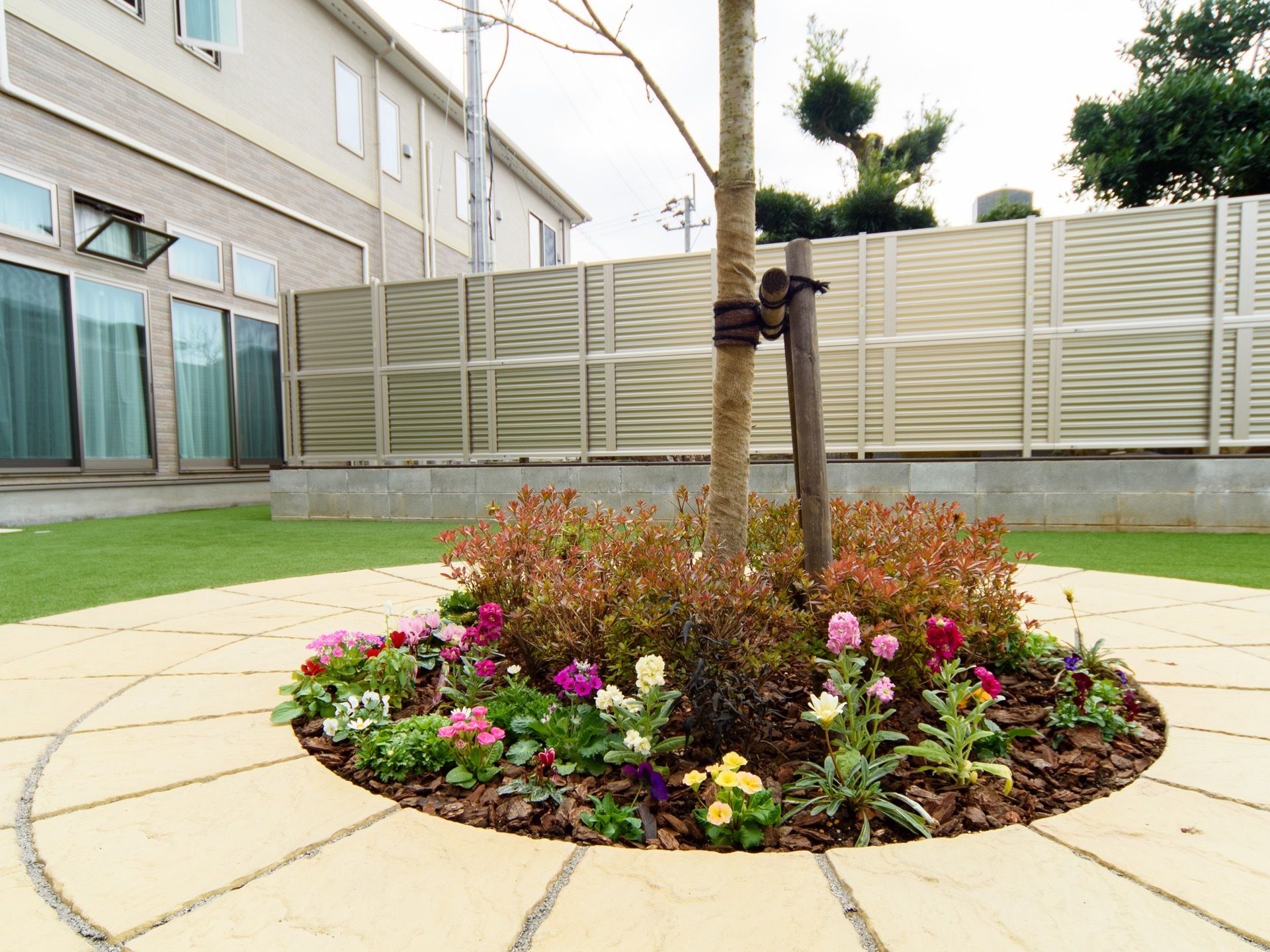 円形に敷かれたレンガの中央に、木が１本植えられている。木の周りには、種類の異なる色とりどりの花が複数ある。