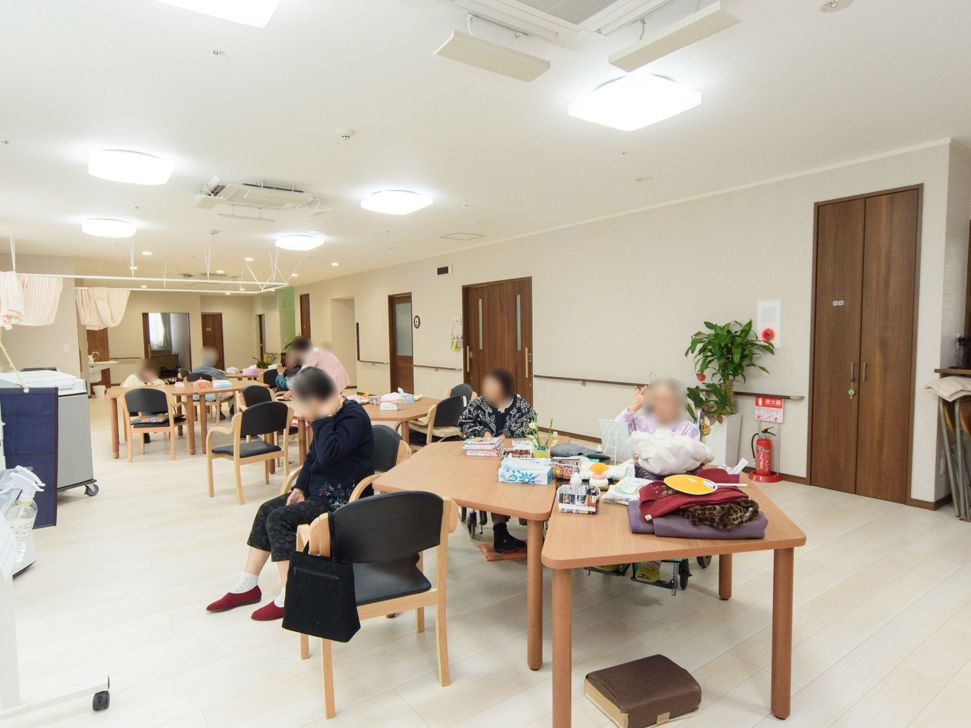 テーブルと椅子が置かれ、複数名の入居者様が椅子に座っている様子である。壁際には観葉植物があり、その横には消火器が設置されている。