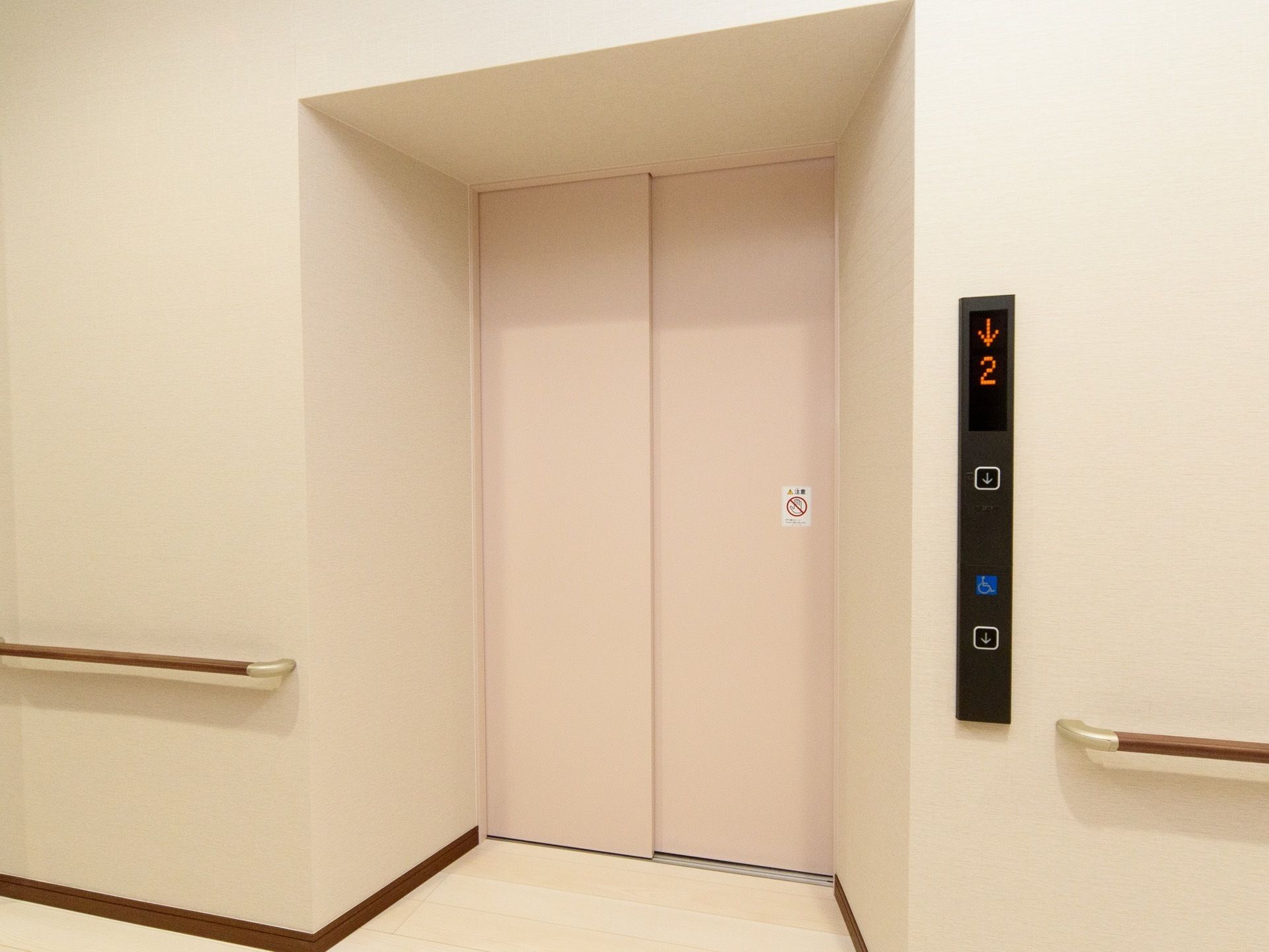 白い壁の間に設置された、１基のエレベーターである。扉は薄いピンク色となっており、壁には木製の手すりが取付けられている。