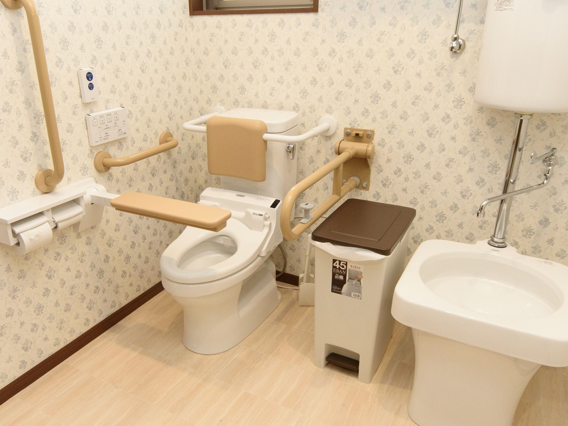 模様の入った壁紙のトイレ空間である。壁には手すりがあり、床には茶色のふたのあるゴミ箱が置かれている。