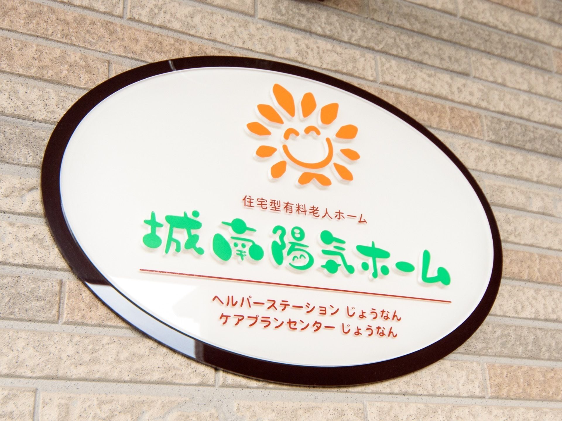 薄茶ロのレンガの外壁に、プレートが取付けられている様子である。楕円形のプレートの中央に、施設名と絵が書かれている。