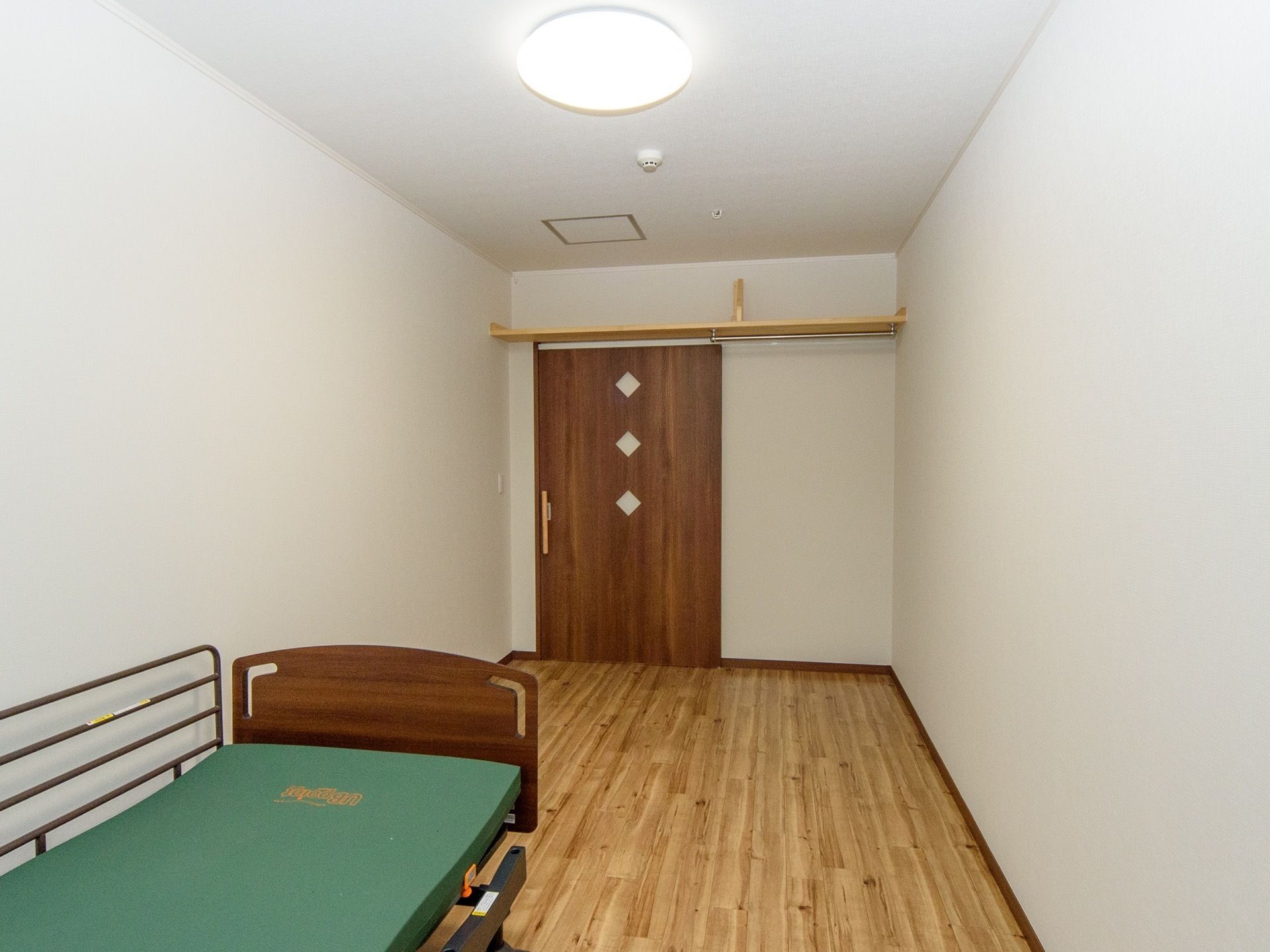 フローリングの床の居室である。ベッドが1台置かれており、マットレスは緑色である。入り口扉上には収納棚が取付けてある。