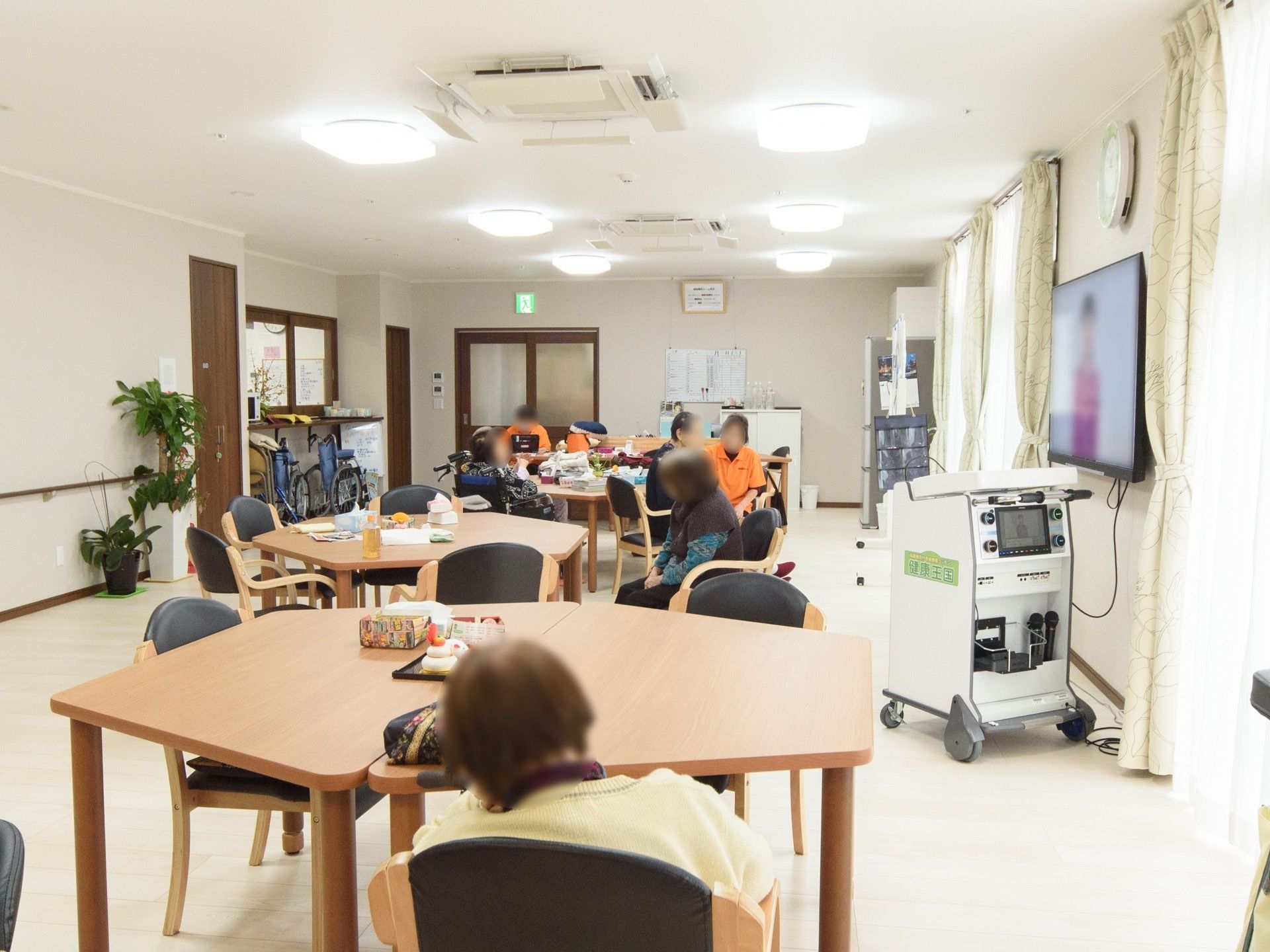六角形のテーブルが、複数並べて置かれており、その周りに入居者様が座っている様子である。壁には、壁掛けテレビが設置されている。