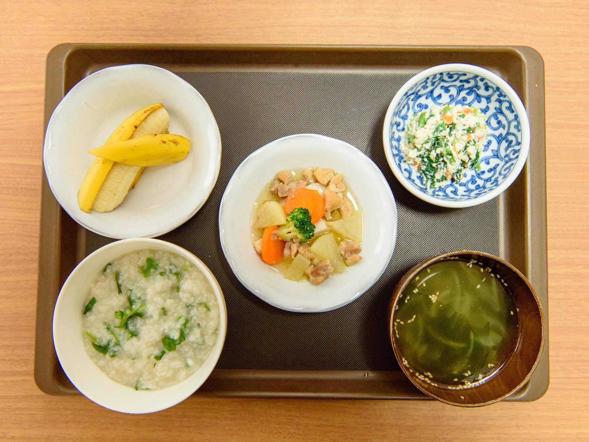 テーブルの上の焦げ茶色のお盆に置かれた、1食分の食事である。野菜の入ったお粥とみそ汁、おかずと果物がある。