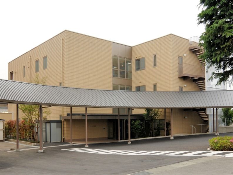 ベージュの３階建ての建物は窓が多く、屋外階段や玄関前に屋根もついている。