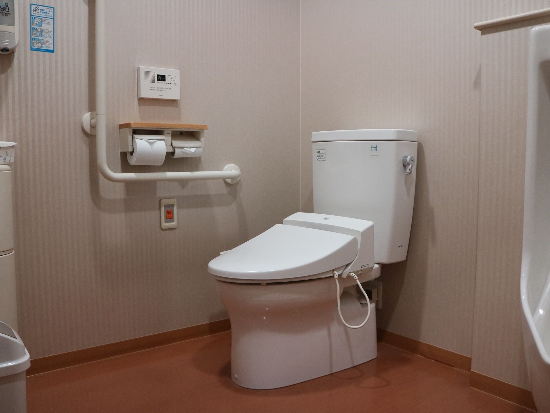 小便器が設けられている個室トイレ。壁にはⅬ字型の手すりやナースコール、２連のペーパーホルダーが設置されている。