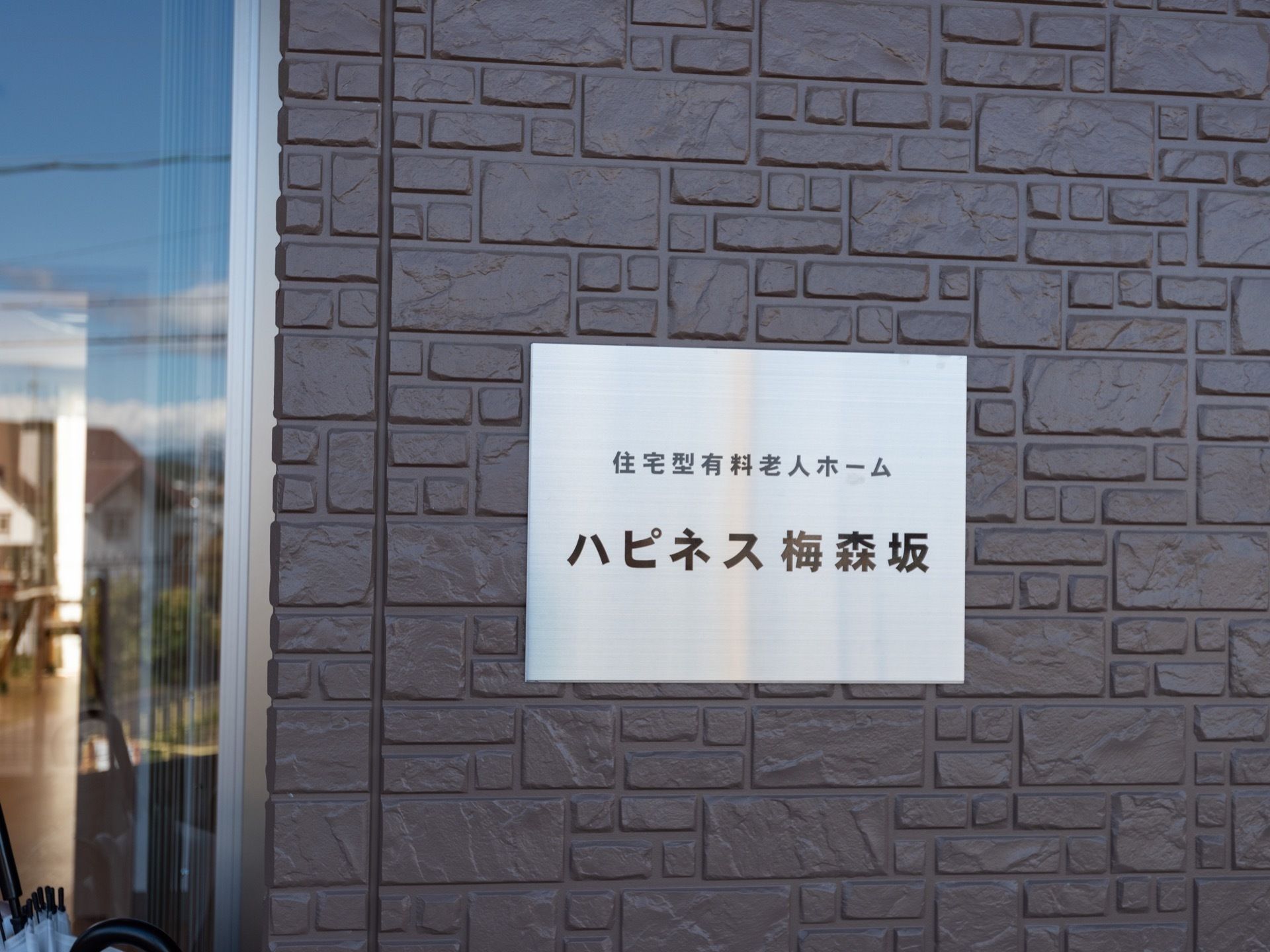 グレーの外壁の上に、施設名が書いてあるラミネート加工された紙が貼られている。外壁の左側はガラス張りで、室内が見える。