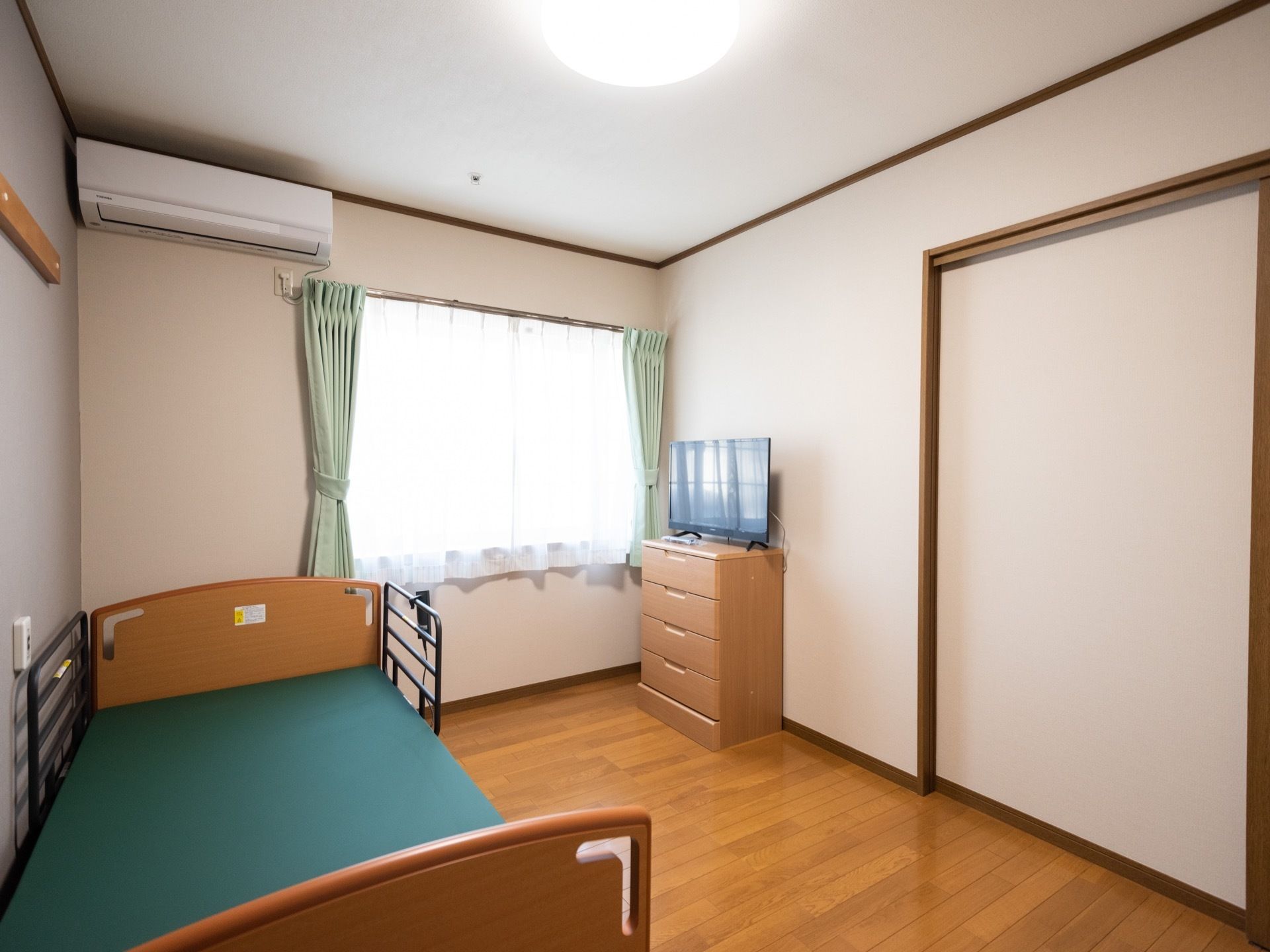 フローリングの部屋にベッドとチェスト、テレビなどが備え付けられている。腰高窓に薄緑色のカーテンが掛けられている。