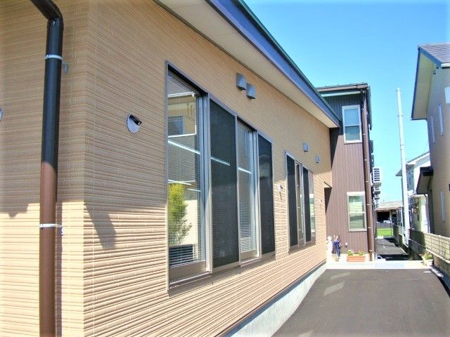 ベージュを基調とした建物の外壁。建物の周辺には段差や通行の邪魔になるようなものがなく、周辺には住宅がある。