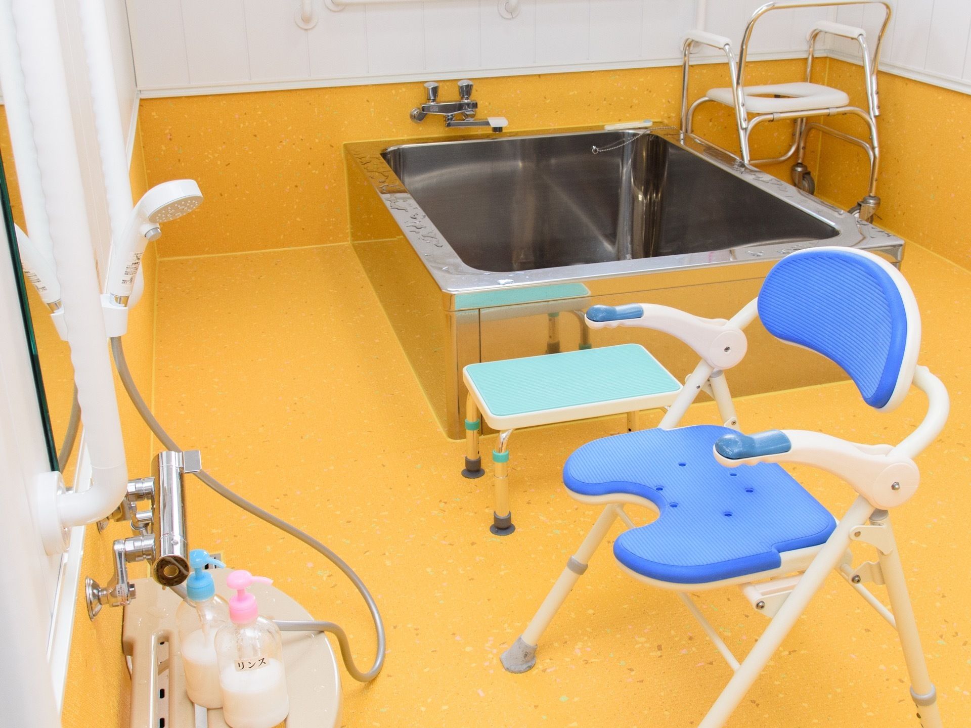 黄色い床の広い浴室がある。洗い場には青いシャワー椅子が置かれており、奥にはステンレスの大きな浴槽が設置されている。
