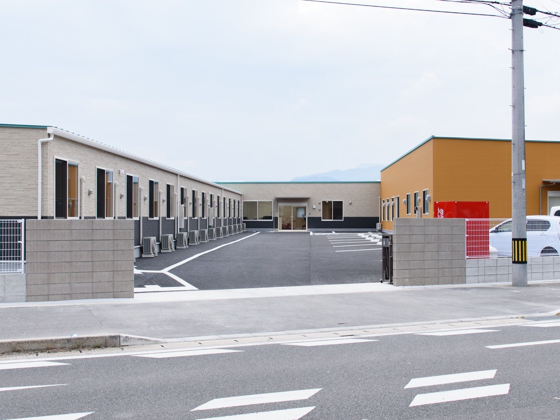 歩道の横に舗装された敷地が広がり、コの字型の平屋建て住宅が建っている。正面には玄関があり、建物の脇は駐車スペースになっている。