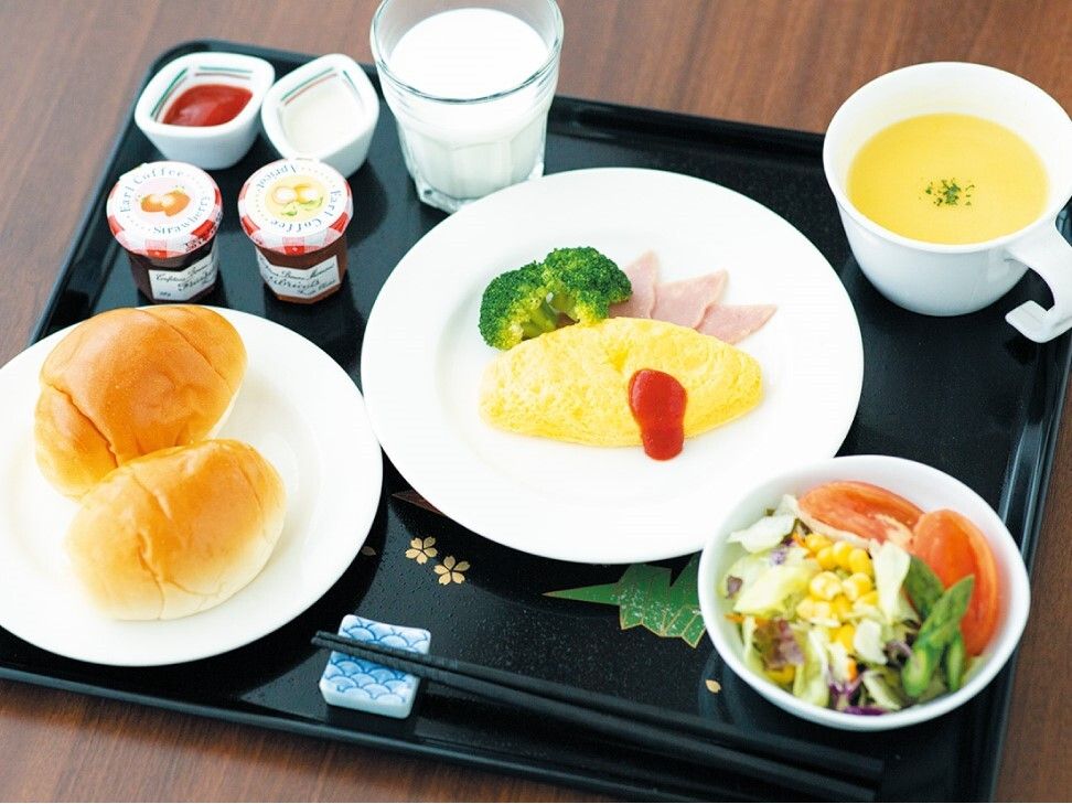 黒いトレーに並べられた食事。ロールパンにオムレツ、サラダ、牛乳やコーンスープなど。