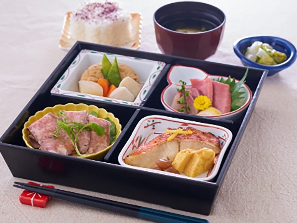 黒いお弁当箱に箸きりが付き、魚料理や肉料理、煮物などがある。その横には吸い物や漬物、ゆかりご飯もある。