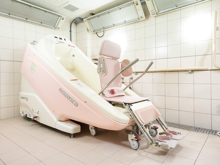 座ったまま入浴可能な機械浴を設置した浴室。白とピンクの車いす風の設備が付いたマシンがある。