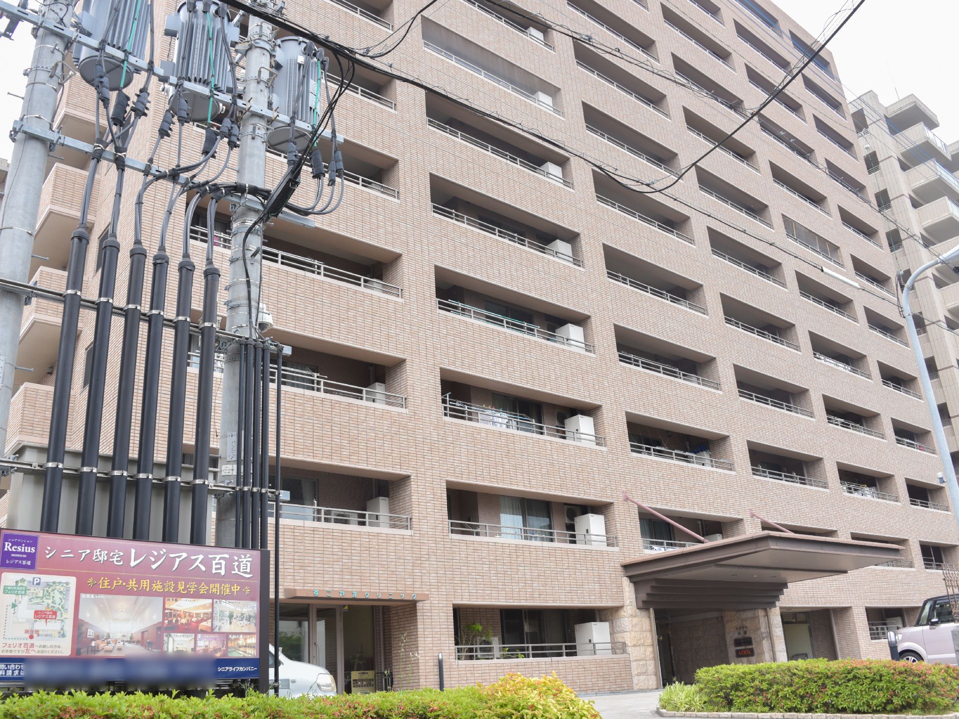 外観は、12階建てのマンションのように見える造りになっている。ブラウンの壁が落ち着いたデザインである。前方には車が置いてある。