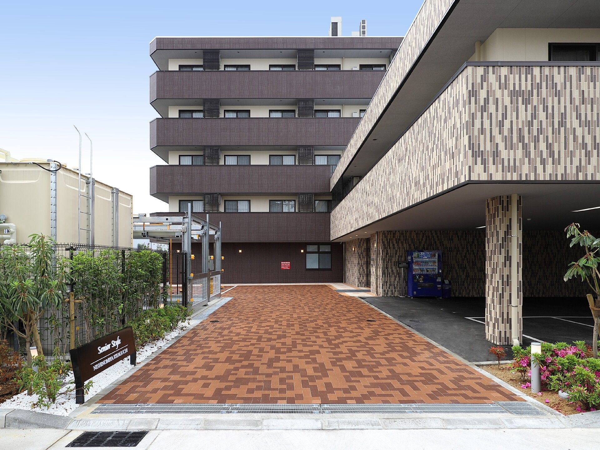 5階建てのダークブラウンの建物が左側にあり、玄関を含む右側の建物は2階建て。駐車スペースも広い。