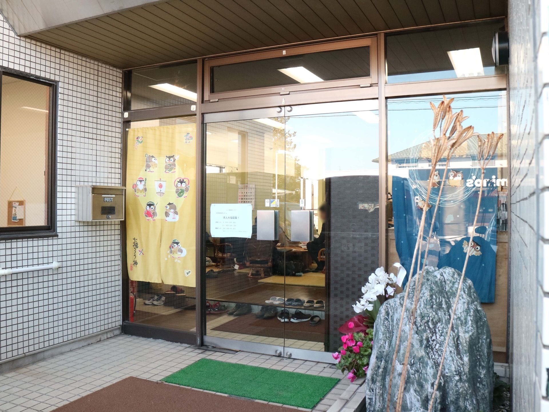 ガラスサッシの両開き扉が設置されている施設入口である。足元には2種類の泥落としマットが敷かれている。